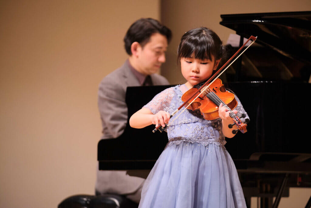 バイオリンの発表会で先生のピアノ伴奏に合わせて、真剣にバイオリンの演奏をする女の子の写真